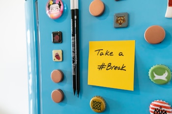 A light blue refrigerator door adorned with various colorful magnets and bottle caps. A bright yellow sticky note is attached with the message 'Take a #Break' written in black ink. Next to the note are two black pens held upright by a magnet.
