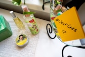 A neatly arranged setup on a white table includes two bottles of spray, one with floral designs, a tin with vintage-style artwork, and a green box. A yellow sticky note with '#SelfCare' and a heart is attached to a mirror. Green leaves add a touch of nature to the scene.