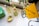 A neatly arranged setup on a white table includes two bottles of spray, one with floral designs, a tin with vintage-style artwork, and a green box. A yellow sticky note with '#SelfCare' and a heart is attached to a mirror. Green leaves add a touch of nature to the scene.