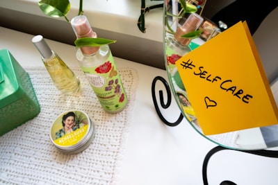 A neatly arranged setup on a white table includes two bottles of spray, one with floral designs, a tin with vintage-style artwork, and a green box. A yellow sticky note with 