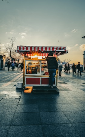 A street vendor cart with a red and white striped canopy is set against a backdrop of a sunset. The sun is setting behind the cart, creating a warm glow through the glass windows. People can be seen walking in the background on a large paved area. The surrounding atmosphere suggests a busy, urban setting, and the overall color palette is warm and inviting with orange and yellow hues.