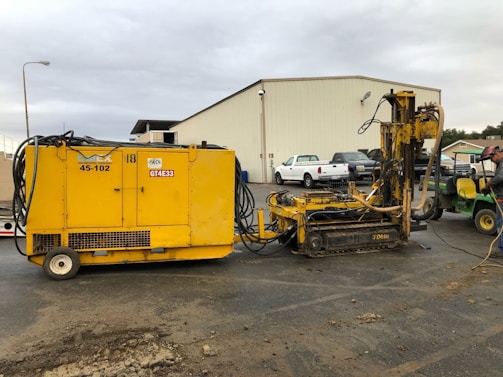 A yellow construction machine with various cables and hoses is positioned on the ground. In the background, there's an industrial building with a corrugated metal exterior. Several vehicles, including a white truck, are parked near the building. A person wearing protective gear is standing beside the machinery, possibly controlling or operating it.