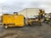 A yellow construction machine with various cables and hoses is positioned on the ground. In the background, there's an industrial building with a corrugated metal exterior. Several vehicles, including a white truck, are parked near the building. A person wearing protective gear is standing beside the machinery, possibly controlling or operating it.