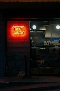 A dimly lit storefront with a bright red neon sign reading 'TAKE AWAY' mounted on a brick wall. Adjacent to it, a small table with a plant and two chairs is set up. Inside the shop, minimalistic black and white decor is visible, including hanging lights and shelving.