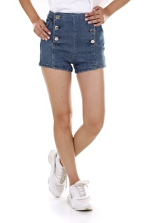 A person is standing with legs crossed wearing high-waisted denim shorts with metallic button details and white sneakers. The casual outfit is paired with a tucked-in white top. The background is plain white, emphasizing the clothing.