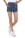A person is standing with legs crossed wearing high-waisted denim shorts with metallic button details and white sneakers. The casual outfit is paired with a tucked-in white top. The background is plain white, emphasizing the clothing.