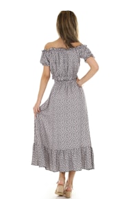 A person with long hair is wearing an off-shoulder, ankle-length floral dress with a ruffle hem. The dress has a subtle pattern in dark tones, and the individual is standing with their back to the camera, holding the dress with one hand. The footwear is wedge sandals with ankle straps.