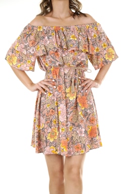 A person is wearing an off-shoulder dress with a floral pattern in warm tones of pink, peach, orange, and yellow. The dress features ruffled layers and a waist tie, and the person has their hands on their hips.