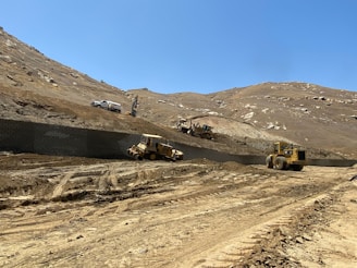 A construction site located on a barren hillside with several heavy machinery vehicles in operation. A pickup truck, a bulldozer, and other construction equipment are visible against a backdrop of dirt and rocky terrain. The sky is clear and blue, indicating a sunny day.