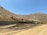 Several large construction vehicles are working on a hilly, dirt-covered terrain. The rugged landscape consists of earth-moving machinery, including excavators, operating on a slope. The sky is clear, suggesting bright sunlight, and the area appears to be a remote or undeveloped natural environment.