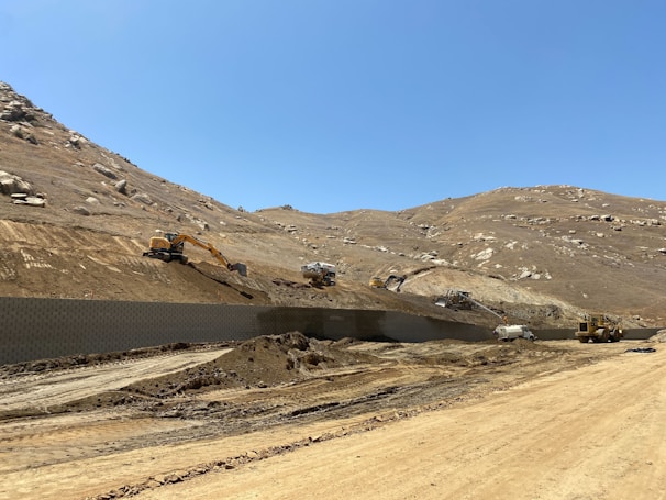 Several large construction vehicles are working on a hilly, dirt-covered terrain. The rugged landscape consists of earth-moving machinery, including excavators, operating on a slope. The sky is clear, suggesting bright sunlight, and the area appears to be a remote or undeveloped natural environment.