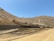 Several large construction vehicles are working on a hilly, dirt-covered terrain. The rugged landscape consists of earth-moving machinery, including excavators, operating on a slope. The sky is clear, suggesting bright sunlight, and the area appears to be a remote or undeveloped natural environment.