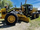 A large yellow motor grader with black and green accents is displayed on a grassy outdoor setting. It features large rubber tires and a prominent front blade. In the background, there are trees, a white tent, and several people, suggesting an outdoor exhibition or machinery event.