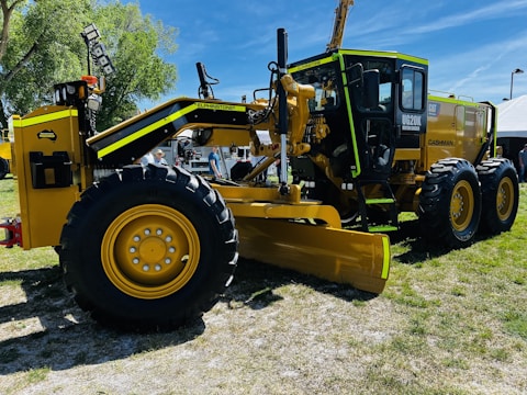 A large yellow motor grader with black and green accents is displayed on a grassy outdoor setting. It features large rubber tires and a prominent front blade. In the background, there are trees, a white tent, and several people, suggesting an outdoor exhibition or machinery event.