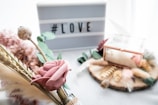 A delicate composition featuring a pink artificial rose surrounded by dried flowers and greenery in the foreground. Behind the floral arrangement, a lightbox sign displays the hashtag LOVE. To the right of the sign, a glass box with a copper frame sits on a wooden slice decorated with feathers and more dried botanicals, enhancing the romantic and rustic aesthetic.