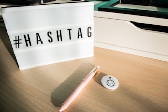 A lightbox with the word hashtag displayed in uppercase letters sits on a wooden desk. A pink pen and a small round pin with a minimalist robot face design are nearby. The background features a white compartment and some shadows, suggesting natural lighting.