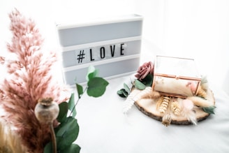 A decorative setup featuring a lightbox with the word '#LOVE' placed on a white background. To the right, there is a glass box with a rose gold frame on a wooden slice decorated with assorted dried flowers and greenery. To the left, there are pink pampas grass and other dried elements, adding a soft, romantic touch.