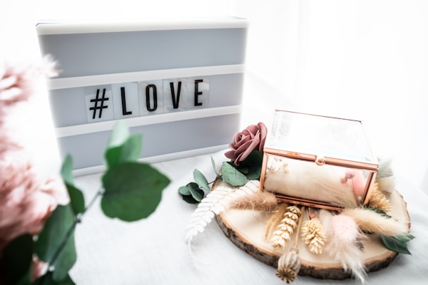 A lightbox with the hashtag 'LOVE' is placed next to a decorative wooden slice adorned with dried flowers, greenery, and a glass box with gold edges. There is a gentle, soft aesthetic with pastel tones and textures from feathers and artificial flowers.