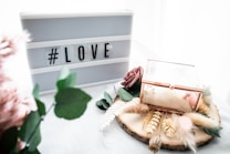 A lightbox with the hashtag 'LOVE' is placed next to a decorative wooden slice adorned with dried flowers, greenery, and a glass box with gold edges. There is a gentle, soft aesthetic with pastel tones and textures from feathers and artificial flowers.