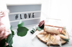 A lightbox with the hashtag 'LOVE' is placed next to a decorative wooden slice adorned with dried flowers, greenery, and a glass box with gold edges. There is a gentle, soft aesthetic with pastel tones and textures from feathers and artificial flowers.