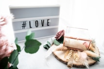 A lightbox with the hashtag 'LOVE' is placed next to a decorative wooden slice adorned with dried flowers, greenery, and a glass box with gold edges. There is a gentle, soft aesthetic with pastel tones and textures from feathers and artificial flowers.