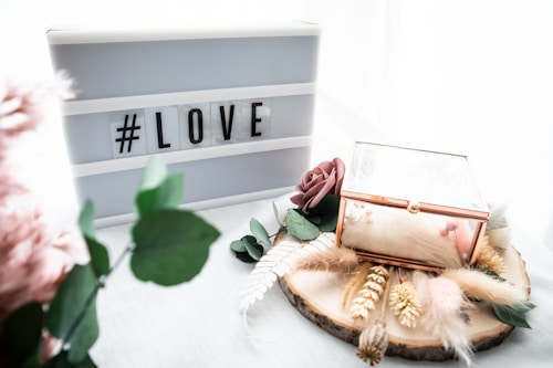 A lightbox with the hashtag 'LOVE' is placed next to a decorative wooden slice adorned with dried flowers, greenery, and a glass box with gold edges. There is a gentle, soft aesthetic with pastel tones and textures from feathers and artificial flowers.