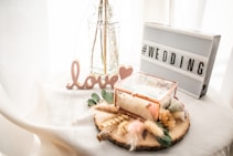 A decorative setup with a glass vase holding dried plants, a lightbox displaying &lsquo;#WEDDING&rsquo;, a wooden love sign, and a glass box containing dried flowers, all on a light-colored draped fabric.