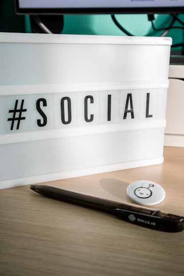 A lightbox sign displaying the word '#SOCIAL' in bold black letters on a desk surface. Next to it, there is a black pen and a round button with a cartoonish face. The background includes a part of a computer monitor and its cords.