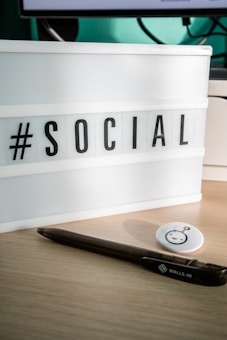 A lightbox sign displaying the word '#SOCIAL' in bold black letters on a desk surface. Next to it, there is a black pen and a round button with a cartoonish face. The background includes a part of a computer monitor and its cords.