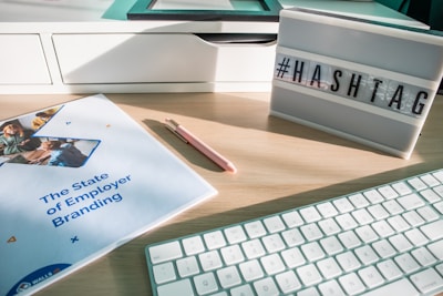 A workspace scene featuring a desk with a white keyboard, a pink pen, a report titled 'The State of Employer Branding', and a lightbox displaying the word '#HASHTAG'. The desk is light wood, and the ambient light creates a bright, modern atmosphere.