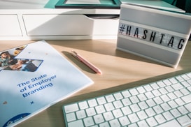 A workspace scene featuring a desk with a white keyboard, a pink pen, a report titled 'The State of Employer Branding', and a lightbox displaying the word '#HASHTAG'. The desk is light wood, and the ambient light creates a bright, modern atmosphere.