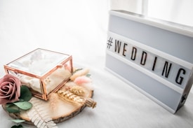 A lightbox with the hashtag 'WEDDING' in bold letters stands on a white surface. Beside it, a small, transparent glass box with a copper frame contains delicate fabric and is adorned with dried flowers and a large artificial rose. The arrangement rests on a round wooden slab, decorated with dried wheat stalks and soft pastel feathers, creating a rustic yet elegant setup.