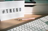 A lightbox displaying the word '#TREND' is placed on a wooden desk. Next to it, a pastel pink pen with golden accents lays on the surface. Part of a white keyboard is visible in the foreground, and there are shadows cast by the afternoon light.
