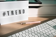 A lightbox displaying the word '#TREND' is placed on a wooden desk. Next to it, a pastel pink pen with golden accents lays on the surface. Part of a white keyboard is visible in the foreground, and there are shadows cast by the afternoon light.
