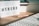 A lightbox displaying the word '#TREND' is placed on a wooden desk. Next to it, a pastel pink pen with golden accents lays on the surface. Part of a white keyboard is visible in the foreground, and there are shadows cast by the afternoon light.