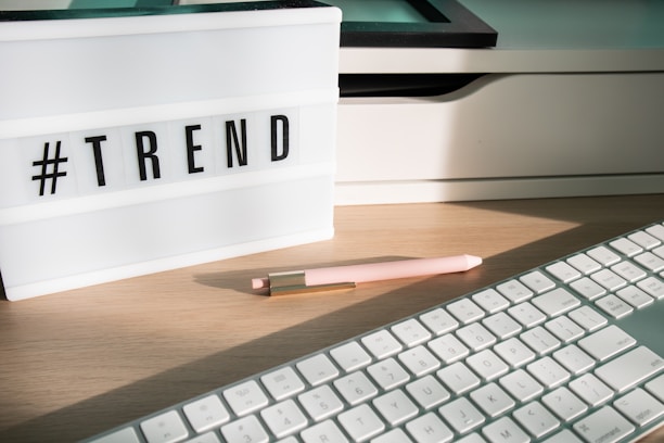 A lightbox displaying the word '#TREND' is placed on a wooden desk. Next to it, a pastel pink pen with golden accents lays on the surface. Part of a white keyboard is visible in the foreground, and there are shadows cast by the afternoon light.