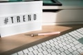 A lightbox displaying the word '#TREND' is placed on a wooden desk. Next to it, a pastel pink pen with golden accents lays on the surface. Part of a white keyboard is visible in the foreground, and there are shadows cast by the afternoon light.