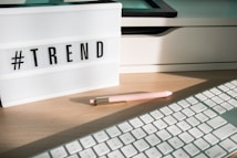 A lightbox displaying the word '#TREND' is placed on a wooden desk. Next to it, a pastel pink pen with golden accents lays on the surface. Part of a white keyboard is visible in the foreground, and there are shadows cast by the afternoon light.