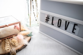 A lightbox displays the word '#LOVE' in bold black letters. Beside it, a wooden slab serves as a base for an arrangement of dried flowers, feathers, and a transparent glass container with a rose gold edge. The setting is airy and softly lit, conveying a cozy and romantic ambiance.