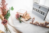 A decorative setup featuring a lightbox with the word 'WEDDING' and a cutout of the word 'love' placed on a white surface. There is a small glass box with a rose, surrounded by dried flowers and greenery. The scene is softened by the presence of delicate pampas grass in a vase.