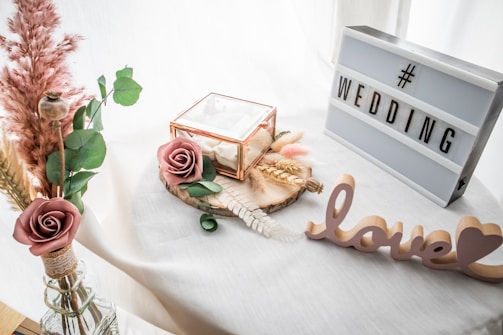 A decorative setup featuring a lightbox with the word 'WEDDING' and a cutout of the word 'love' placed on a white surface. There is a small glass box with a rose, surrounded by dried flowers and greenery. The scene is softened by the presence of delicate pampas grass in a vase.