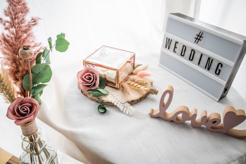 A decorative setup featuring a lightbox with the word 'WEDDING' and a cutout of the word 'love' placed on a white surface. There is a small glass box with a rose, surrounded by dried flowers and greenery. The scene is softened by the presence of delicate pampas grass in a vase.