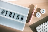 A lightbox displaying the word '#TREND' is placed on a wooden desk. Nearby, there's a pink pen with a gold clip, two round badges with icons, and part of a computer keyboard.