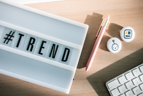 A lightbox displaying the word '#TREND' is placed on a wooden desk. Nearby, there's a pink pen with a gold clip, two round badges with icons, and part of a computer keyboard.