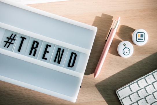 A lightbox displaying the word '#TREND' is placed on a wooden desk. Nearby, there's a pink pen with a gold clip, two round badges with icons, and part of a computer keyboard.