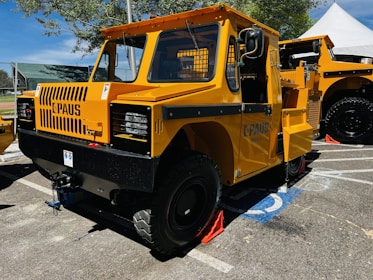 A bright yellow industrial vehicle with a rugged design, featuring large black tires, a boxy cabin, and grill on the front. The vehicle has branding on the side and is parked in an outdoor area with a blue sky and trees in the background. There are traffic cones placed around it.