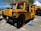 A bright yellow industrial vehicle with a rugged design, featuring large black tires, a boxy cabin, and grill on the front. The vehicle has branding on the side and is parked in an outdoor area with a blue sky and trees in the background. There are traffic cones placed around it.