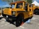 A bright yellow industrial vehicle with a rugged design, featuring large black tires, a boxy cabin, and grill on the front. The vehicle has branding on the side and is parked in an outdoor area with a blue sky and trees in the background. There are traffic cones placed around it.