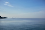 A tranquil ocean scene with calm blue water stretching to the horizon. The sky is clear with a few scattered clouds, and a distant coastline is visible on the left side. The atmosphere conveys a sense of peace and serenity.