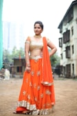 A woman is standing confidently outdoors, wearing a traditional Indian lehenga choli. The outfit is bright orange with intricate gold embroidery and detailing. She accessorizes with gold jewelry, including a choker necklace and bangles. The background features an urban setting with buildings and greenery.